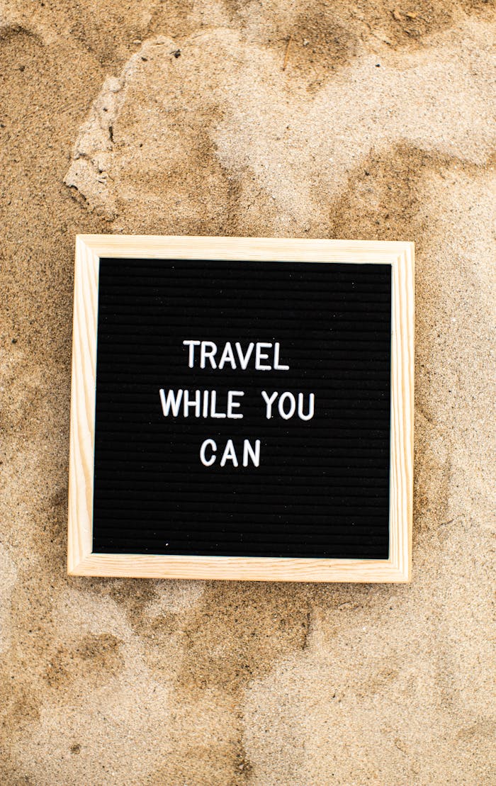 Wooden-framed message board on a sandy beach with text 'Travel While You Can'.